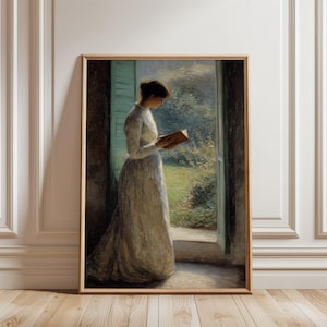Puede incluir: Una obra de arte enmarcada representa a una mujer con un vestido largo blanco leyendo un libro junto a una ventana. La obra tiene una estética vintage, con colores suaves y un enfoque en la luz natural. La mujer está de pie frente a una ventana con contraventanas verdes, mirando un jardín.