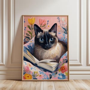 May include: A framed art print featuring a Siamese cat with blue eyes, resting on a floral pillow and reading a book. The background is a soft pink with colorful flowers. The artwork is in a wooden frame.