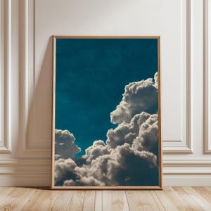 May include: Framed art print featuring a dramatic cloudscape. The artwork showcases fluffy, white clouds against a deep teal sky. The print is encased in a simple, light-toned wooden frame, adding a touch of natural warmth to the scene.