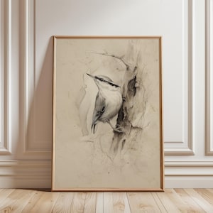 May include: A framed pencil sketch depicting a bird perched on a tree branch. The bird has a white breast and grey wings and back. The artwork is in a wooden frame and leans against a white wall and wooden floor.
