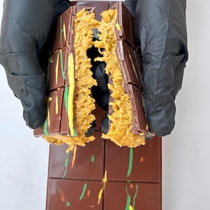 May include: A chocolate bar with a caramel filling, broken in half to reveal the filling. The chocolate is dark brown with green and yellow swirls. The caramel is a light brown colour.