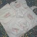 May include: A white baby clothing set featuring a gown, blanket, bib, and hat. Each item is embroidered with a pink bow and floral design, along with the name "Ellie Rae" in pink script. The set has pink trim.