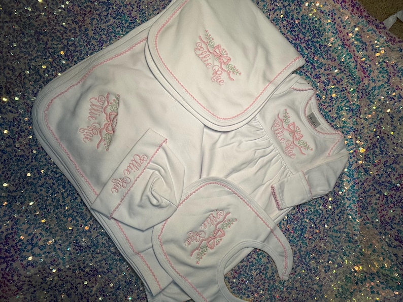 May include: A white baby clothing set with pink embroidered details. The set includes a blanket, bib, hat, and long-sleeved shirt. Each item features the name "Ellie Rae" and a bow design.