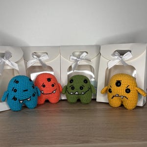 Può includere: Quattro giocattoli mostro all'uncinetto colorati in blu, arancione, verde e giallo, ognuno con occhi neri e una bocca cucita. Sono esposti su una mensola di legno, con scatole regalo bianche legate con nastri sullo sfondo.