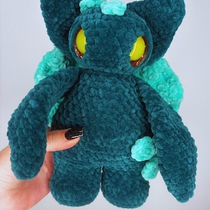 Könnte beinhalten: Ein tealfarbenes Plüschtier mit großen gelben Augen und kleinen tealfarbenen Akzenten. Das handgefertigte Stofftier hat große Ohren und einen kleinen Büschel Teal auf dem Kopf. Die Kreatur wird von einer Hand mit einer schwarzen Maniküre gehalten.