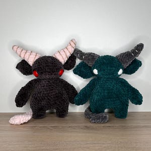 Op de afbeelding: Twee pluche monsterpoppen. Eén is donkergrijs met roze gekleurde hoorns en rode ogen, de andere is blauwgroen met grijze hoorns en witte ogen. Beide hebben kleine armen, benen en staarten. Ze zijn handgemaakt en zitten op een houten oppervlak.