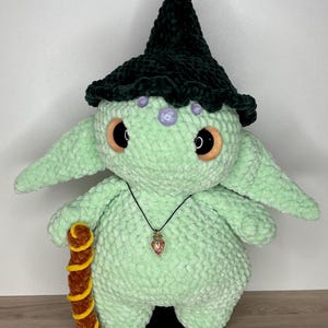 Könnte beinhalten: Eine mintgrüne gehäkelte Puppe mit großen, orangefarben umrandeten Augen und einem dunkelgrünen Hexenhut. Die Puppe hat eine kleine Anhängerkette und hält einen braunen und gelben Stab. Der Hut der Puppe ist mit lila Perlen verziert.