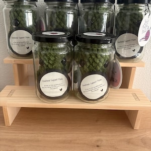 Op de afbeelding: Verschillende glazen potten met zwarte deksels, elk met een groen, getextureerd item. De potten zijn gerangschikt op een kleine houten plank. Elke pot heeft een label met "Emotional Support Pickle."