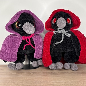 Op de afbeelding: Twee zwarte pluche vogel figuren met grijze snavels en poten. Eén draagt een paarse capuchon cape vastgebonden met roze garen, de andere een rode cape vastgebonden met grijs garen. De vogels hebben rode ogen en staan op een houten oppervlak.