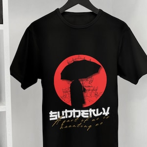 Puede incluir: Una camiseta negra de manga corta con un gráfico de una persona sosteniendo un paraguas sobre un fondo circular rojo con el contorno de un edificio. El texto dice "SUDDENLY" en blanco y "A part of me is haunting me" en dorado.