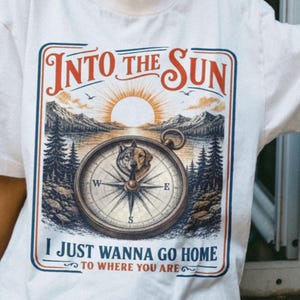 Puede incluir: Camiseta blanca con un gráfico que presenta una brújula con un lobo, montañas y un sol. El texto dice "Into the Sun" y "I Just Wanna Go Home To Where You Are". El diseño tiene una estética vintage y de exteriores.
