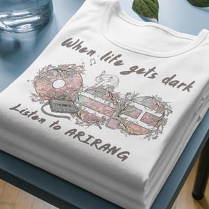 Puede incluir: Camiseta blanca con un diseño gráfico que presenta un disco, una cinta de cassette y un altavoz. El texto "When life gets dark, Listen to ARIRANG" está impreso en la camiseta con una fuente estilizada. La camiseta está doblada y apilada.