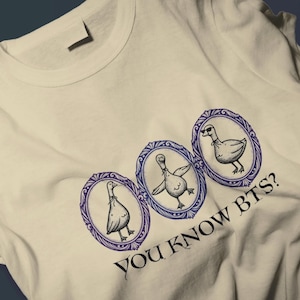 Pode incluir: Camiseta branca com um desenho de três gansos de desenho animado em molduras ovais azuis e roxas. Os gansos são ilustrados em um estilo lúdico, com um usando óculos de sol. O texto "YOU KNOW BTS?" é impresso abaixo.