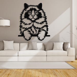 May include: A black metal cat wall art piece. The cat has fluffy fur and is perched on a horizontal bar. The cat's face is detailed with eyes, nose, and whiskers. The art is mounted on a neutral-colored wall above a white sofa.
