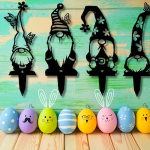 May include: Four black metal garden gnome silhouettes with flowers and butterflies. The gnomes are standing on stakes. The stakes are inserted into a wooden surface. There are seven colorful Easter eggs in the foreground.