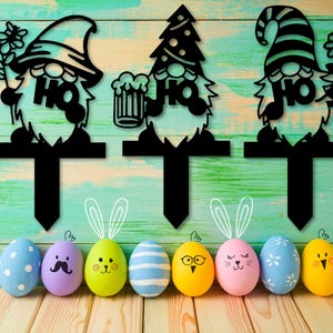 Könnte beinhalten: Drei schwarze Silhouette-Zwergen-Kuchentopper mit einem Bierkrug, einem Weihnachtsbaum und einer Blume. Die Zwerge tragen Hüte und haben Bärte. Die Topper werden auf einem Holzgrund mit bunten Ostereiern gezeigt.