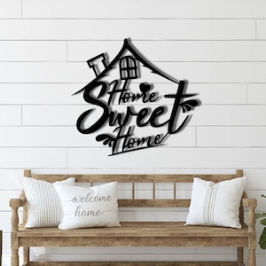 Puede incluir: Arte de pared de metal negro con forma de casa con las palabras "Home Sweet Home" en fuente cursiva. El arte está montado en una pared de tablones horizontales blancos. Un banco de madera con cojines está en primer plano.