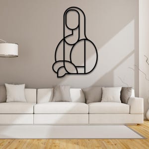 Op de afbeelding: Zwarte metalen wandkunst die een geometrische interpretatie van de Mona Lisa weergeeft. Het kunstwerk is gemonteerd op een witte muur boven een witte bank.