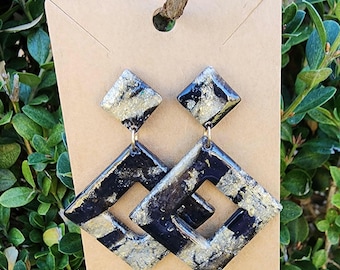 May include: A pair of black and gold geometric dangle earrings. The earrings are made of resin and have a square shape with a cutout in the center. The earrings are accented with gold glitter.