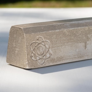 May include: A rectangular concrete block with a trapezoidal profile, featuring a debossed atom symbol on the front face. The surface has a natural, textured gray finish with subtle variations in tone and a small social media handle at the bottom.