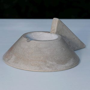 May include: A light gray, conical concrete ashtray with a smooth, matte finish. The object features a wide, circular base that tapers upward to a central bowl with a small notch on the rim. A triangular fin extends from one side of the bowl.
