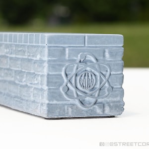 May include: A light gray rectangular brick-textured block featuring a raised atomic symbol emblem on the side. The surface has a matte finish with visible mortar lines between the bricks. The atomic icon consists of three intersecting orbital rings surrounding a central circular core with vertical lines.