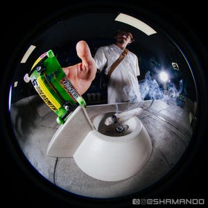 May include: A bright green fingerboard with the text GOLDSMITH and DOKEND printed on the deck, featuring black trucks and neon green wheels, positioned next to a white ceramic ashtray containing a lit, smoking cigarette with visible gray ash.