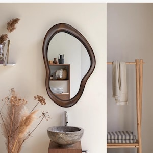 May include: A brown wooden mirror with an organic, free-form shape hangs on a white wall above a stone sink. The mirror reflects a shelf with decorative items.