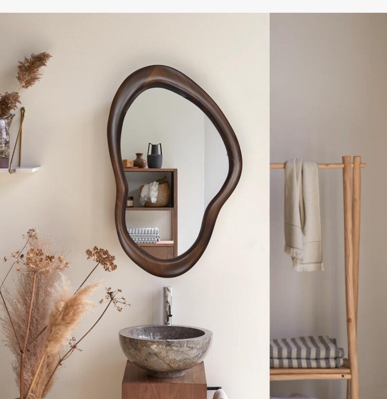 May include: A brown wooden mirror with an organic, free-form shape hangs on a white wall above a stone sink. The mirror reflects a shelf with decorative items.
