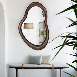 May include: A wooden bench with a light blue cushion and two pillows, a brown wooden frame, and a brown leather strap. A large, irregularly shaped, brown wooden framed mirror hangs on the wall behind the bench.