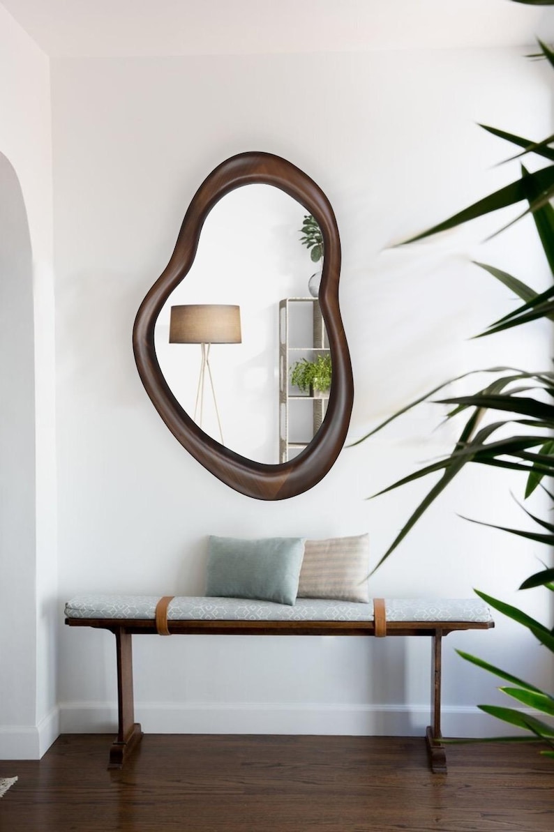 May include: A wooden bench with a light blue cushion and two pillows, a brown wooden frame, and a brown leather strap. A large, irregularly shaped, brown wooden framed mirror hangs on the wall behind the bench.