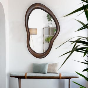 May include: A wooden bench with a light blue cushion and two pillows. The bench has a brown leather strap across the front. A large, dark brown, organic-shaped mirror hangs on the wall behind the bench.