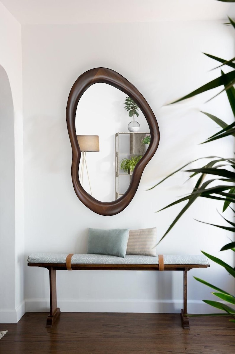 May include: A wooden bench with a light blue cushion and two pillows. The bench has a brown leather strap across the front. A large, dark brown, organic-shaped mirror hangs on the wall behind the bench.
