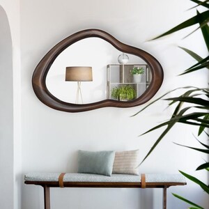 May include: A wooden bench with a light blue cushion and two pillows, one light blue and one beige. The bench is in front of a white wall with a large, dark brown, organic-shaped mirror hanging above it. The mirror reflects a lamp and a small shelf with plants.