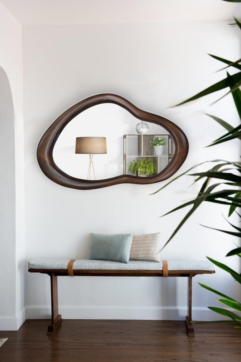 May include: A wooden bench with a light blue cushion and two pillows, one light blue and one beige. The bench is in front of a white wall with a large, dark brown, organic-shaped mirror hanging above it. The mirror reflects a lamp and a small shelf with plants.