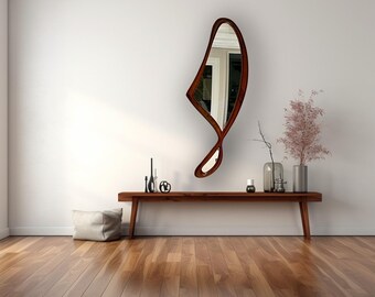 Puede incluir: Un espejo de pared moderno de forma abstracta con un marco de madera marrón. El espejo está colgado en una pared blanca sobre una consola de madera con un jarrón decorativo y velas.