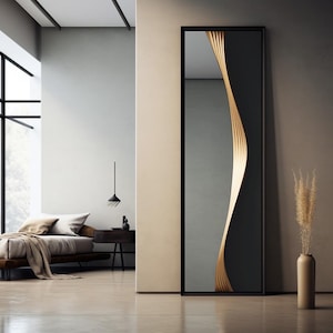 May include: A tall, rectangular mirror with a black frame. A vertical, wavy design of golden wood slats adorns the right side of the mirror. The mirror is in a modern bedroom setting with a bed and a vase with dried plants.