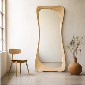 May include: A full-length, free-standing mirror with a unique, wavy wooden frame. The mirror is tall and slender, with a light wood finish. A wooden chair and a terracotta vase with dried branches are also in the room.