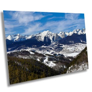 Pode incluir: Uma vista panorâmica de uma cordilheira coberta de neve com um vale em primeiro plano. As montanhas estão cobertas de neve e o vale está coberto de árvores. O céu é azul com nuvens brancas.