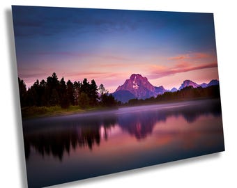 Puede incluir: Una fotografía de paisaje que presenta una cordillera reflejada en un lago tranquilo al atardecer. El cielo es de un rosa y morado vibrantes, con nubes que proyectan largas sombras sobre el agua. Una línea de árboles bordea el lago en primer plano.