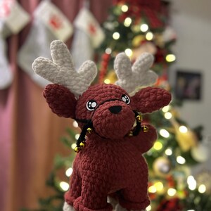 May include: A handmade, plush reindeer toy in a deep red color with cream-colored antlers and hooves. The reindeer has black eyes and a black nose, with gold-colored bells around its neck. The background includes a Christmas tree and stockings.