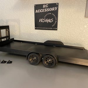 May include: A black RC trailer with two axles and two wheels. The trailer has a flatbed design with a ramp at the rear. The trailer is next to a black RC truck. The sign in the background says "RC Accessory Designs".