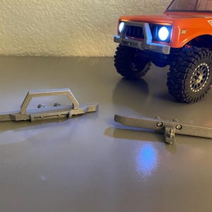 May include: Two gray metal bumpers for a model RC truck. One bumper has a rectangular shape with a handle, and the other is a straight bar with mounting hardware. An orange RC truck with black tires is in the background.