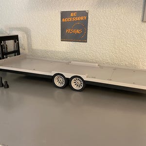 May include: A model RC car trailer with a flatbed design. The trailer has a light gray surface, black trim, and white wheels. The trailer is designed to transport RC vehicles. The sign in the background says "RC Accessory Designs".