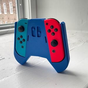 May include: A blue 3D-printed controller grip mount holding a cyan left Joy-Con and a red right Joy-Con. The grip features a textured plastic surface with cutouts for the controller buttons, joysticks, and triggers, providing a secure housing for the gaming device.