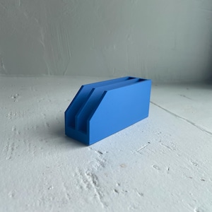 Op de afbeelding: Een helderblauwe bureau-organizer met drie compartimenten. De organizer is gemaakt van een solide materiaal en heeft een eenvoudig, modern ontwerp. Hij staat op een wit oppervlak.