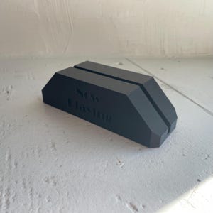 May include: A matte black rectangular display stand featuring a beveled top edge and a central vertical slot for holding items. The front face of the stand is embossed with the text "NOW PLAYING" in a clean, sans-serif font style.