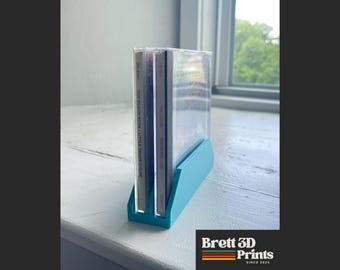 May include: A bright turquoise plastic stand holding two compact disc cases upright. The stand features a rectangular base with two vertical slots designed to secure the cases. The spines of the CDs are visible, displaying text including The Beach Boys.