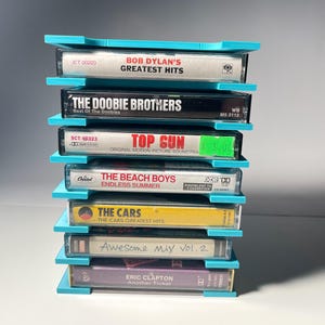 Può includere: Una pila di sette musicassette, ciascuna etichettata con l'artista e il titolo dell'album. I nastri sono tenuti in posizione da un supporto per musicassette azzurro. I titoli includono "Bob Dylan's Greatest Hits", "The Doobie Brothers" e "Top Gun".