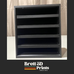 Puede incluir: Un organizador de almacenamiento rectangular negro con cuatro estantes horizontales que crean cinco compartimentos abiertos. Debajo del organizador, el texto dice Brett 3D Prints con un gráfico de rayas horizontales en colores naranja, verde y amarillo.